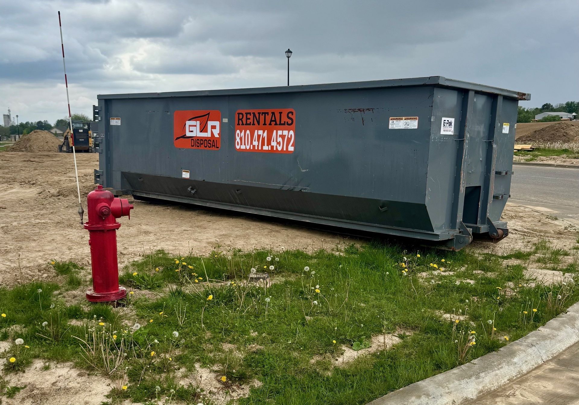 A grey GLR dumpster parked on a dirt lot next to a red fire hydrant under a cloudy sky.