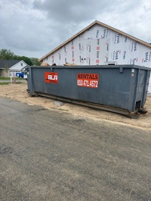 A large grey dumpster from GLR Rentals sits on a dirt patch in front of a house under construction covered in Tyvek wrap.