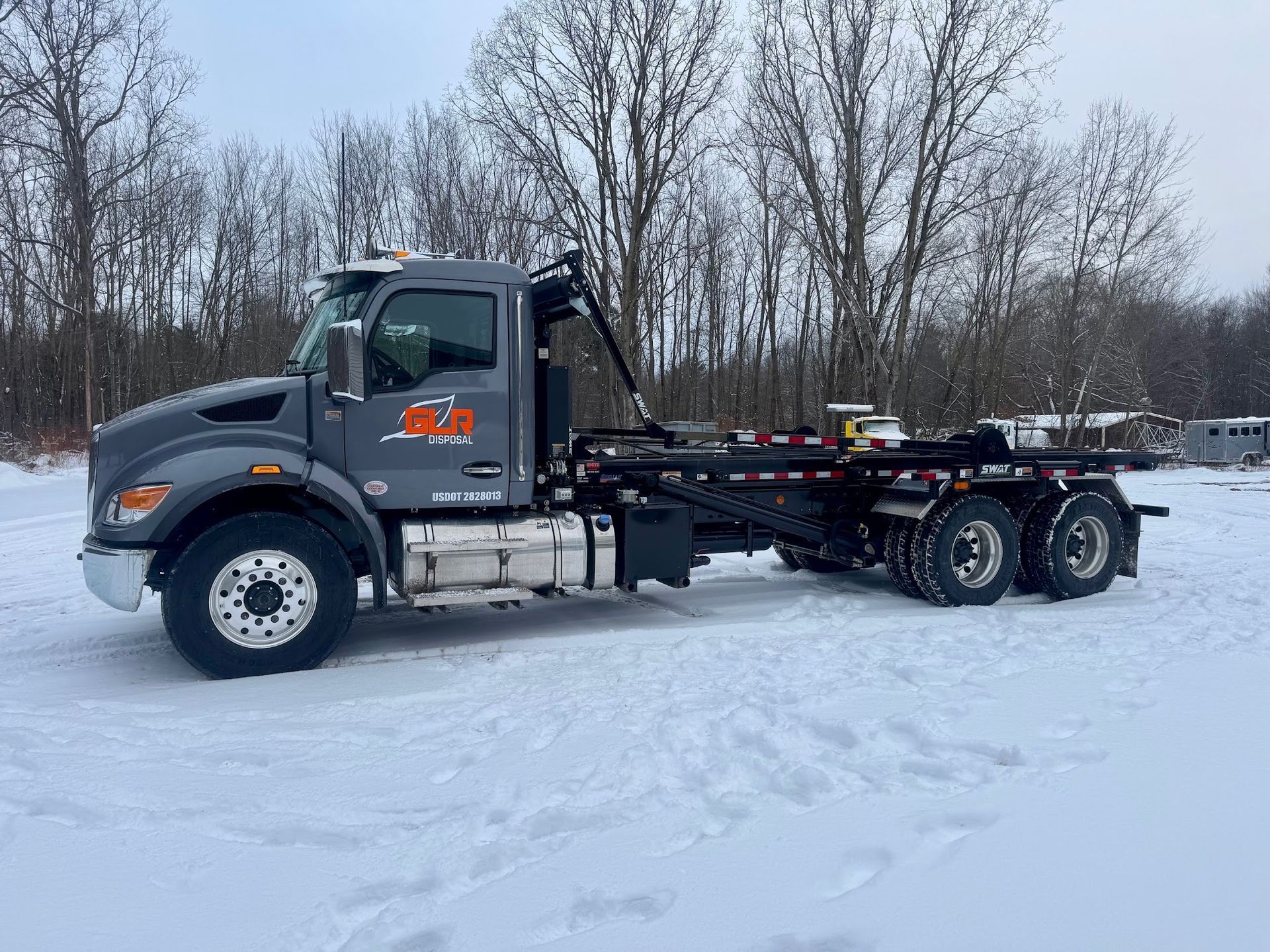 A gray roll-off dump truck parked on a snow-covered field with a treeline in the background.