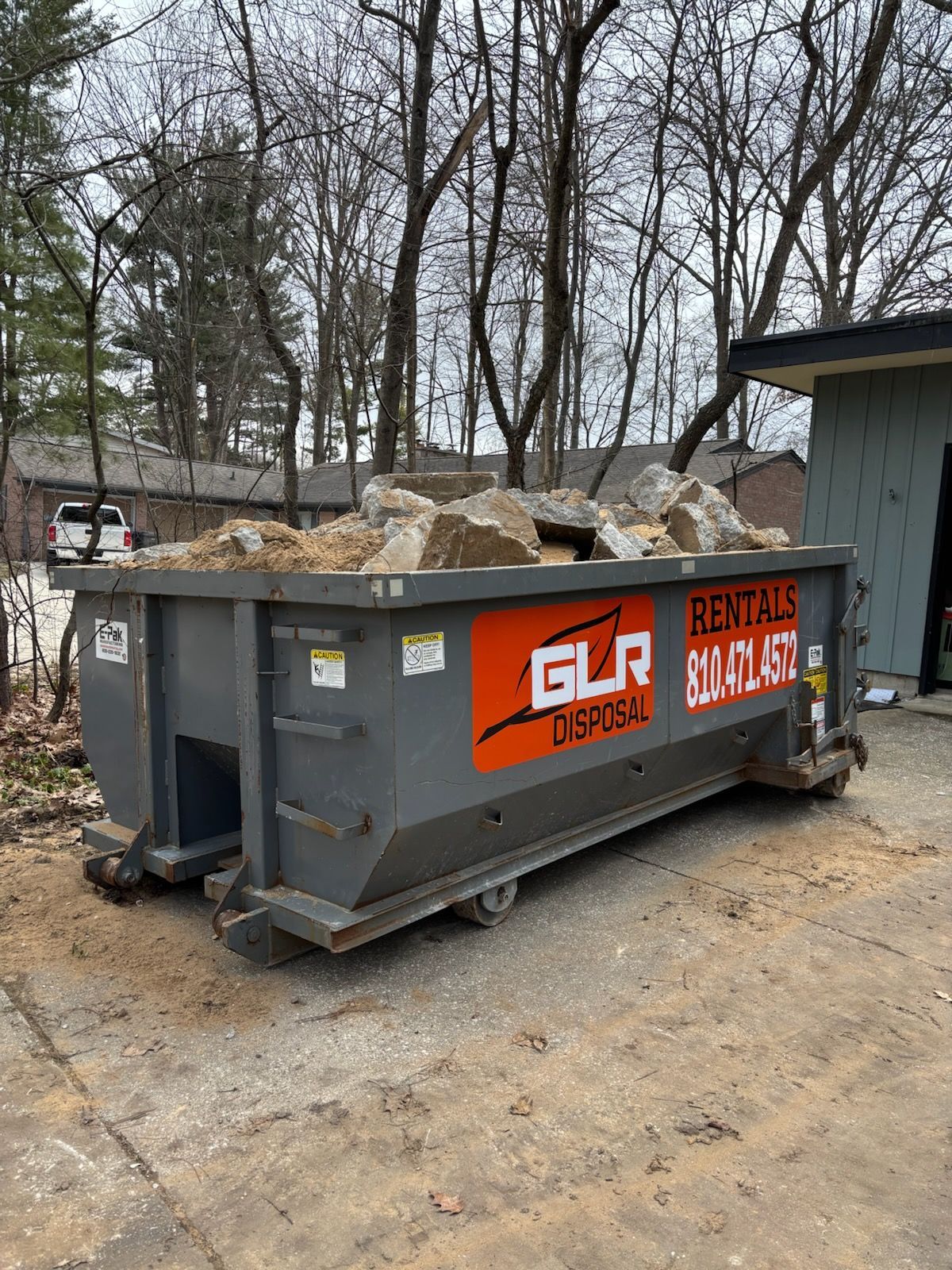 A gray GLR Disposal roll-off dumpster filled with large rocks sits on a concrete surface in front of bare trees and a shed.