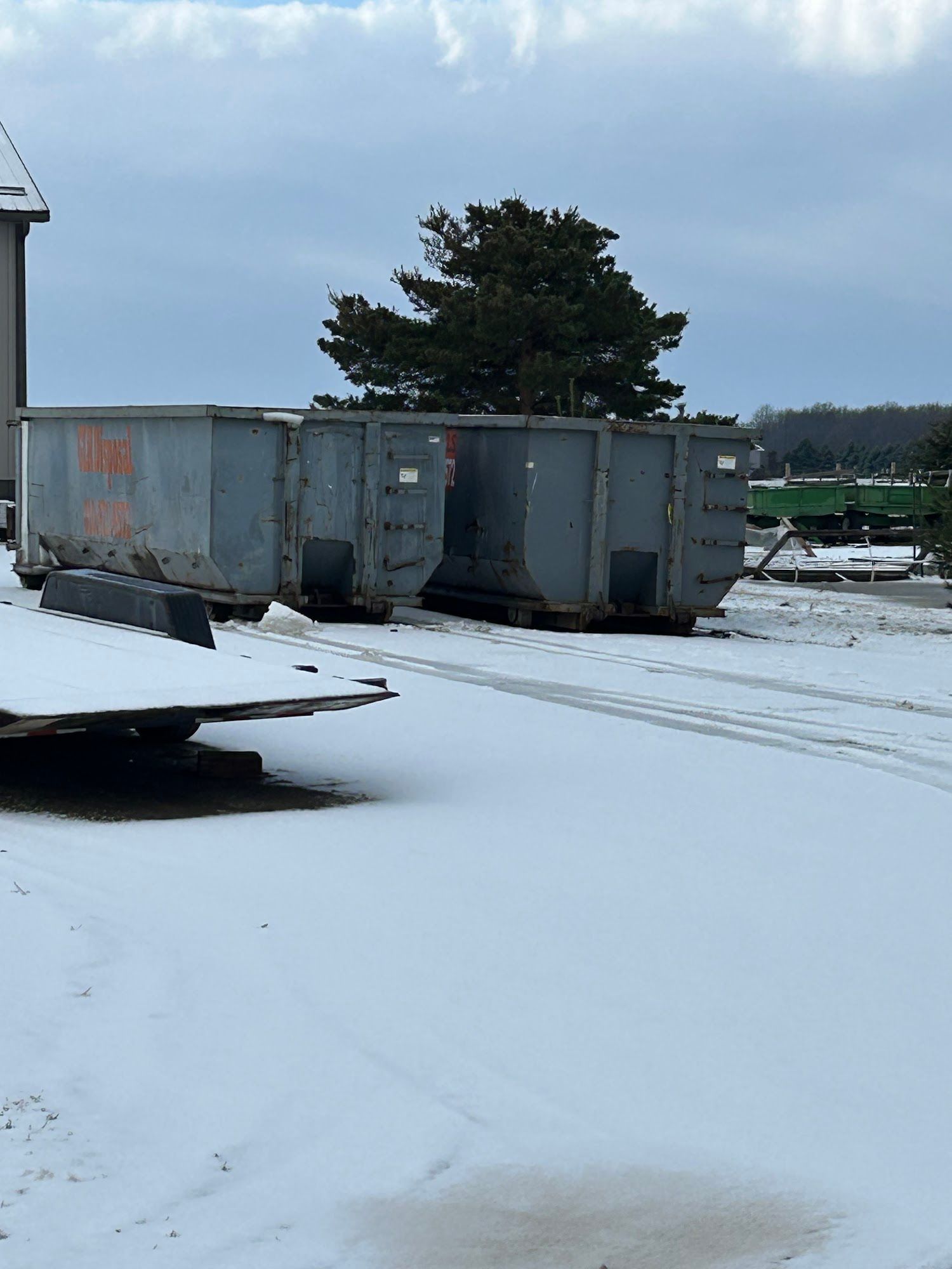 Two large, grey metal industrial dumpsters sit outside in a snowy field with a tree in the background.