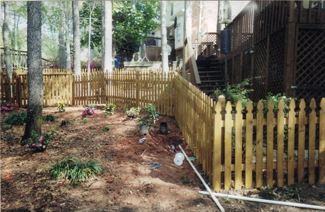 A wooden picket fence surrounds a yard with a house in the background