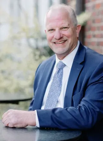 Smiling older man in blue suit, seated outdoors by a brick wall.