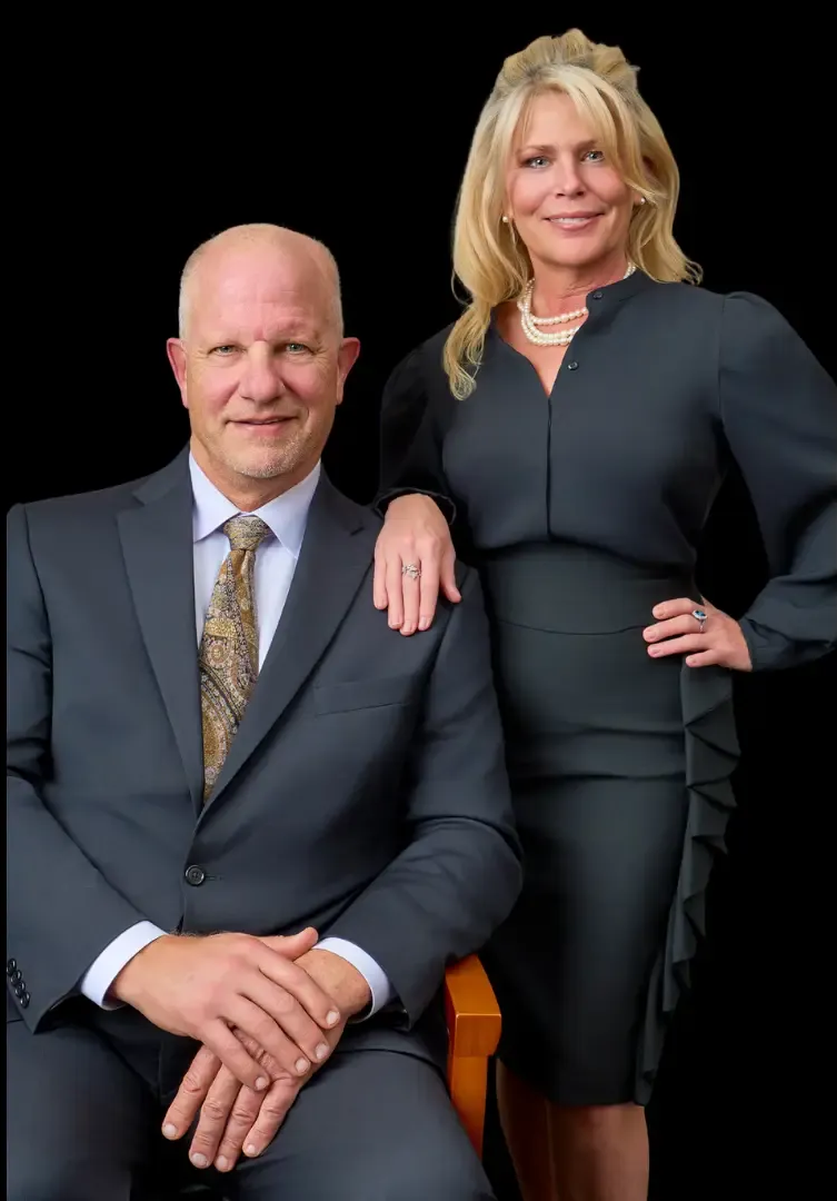 Woman and man sit at a table outside. The woman wears white, the man a blue suit; both smile.