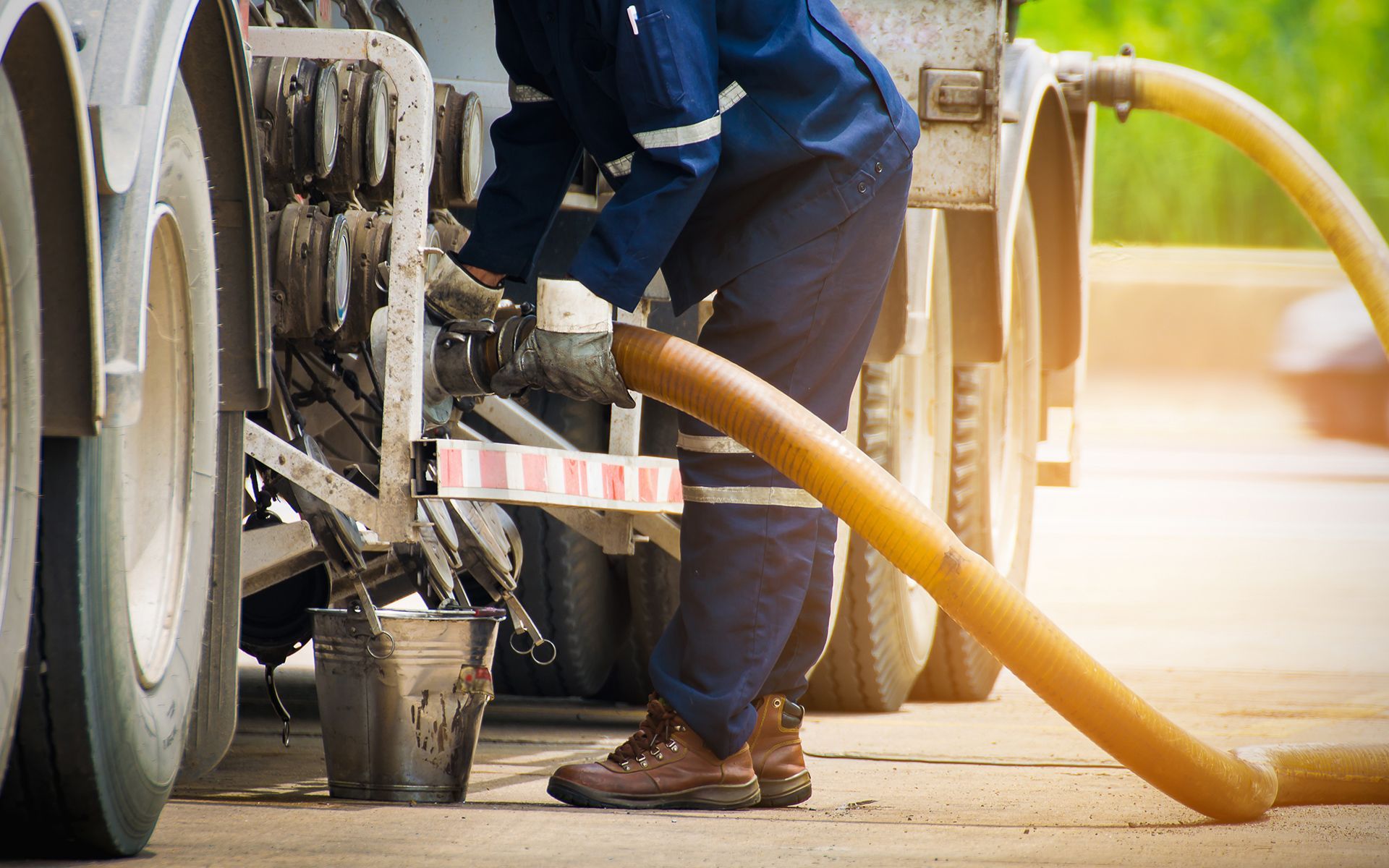 Worker connecting hose to tanker truck, pouring into bucket on a sunny day.