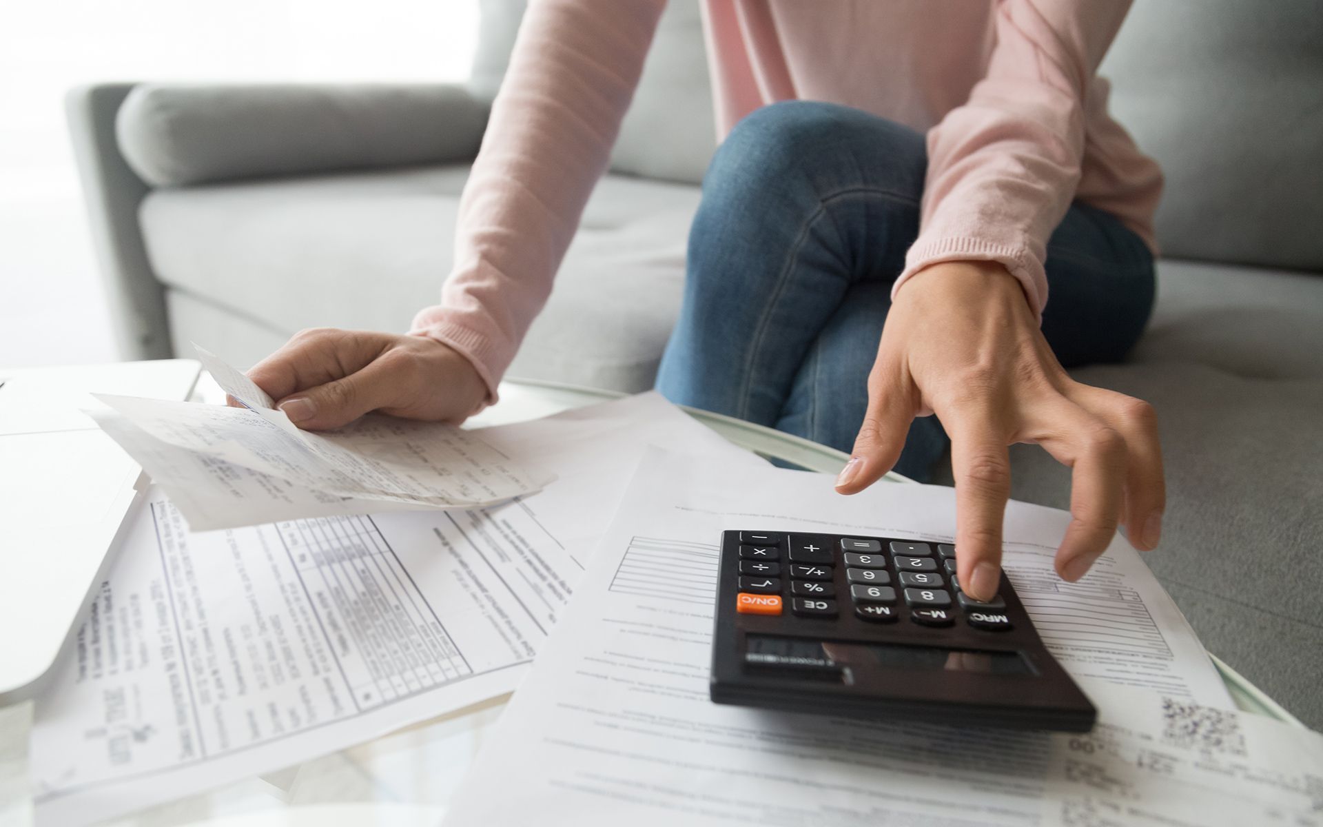 Woman on a couch uses a calculator with bills scattered around, showing financial strain.