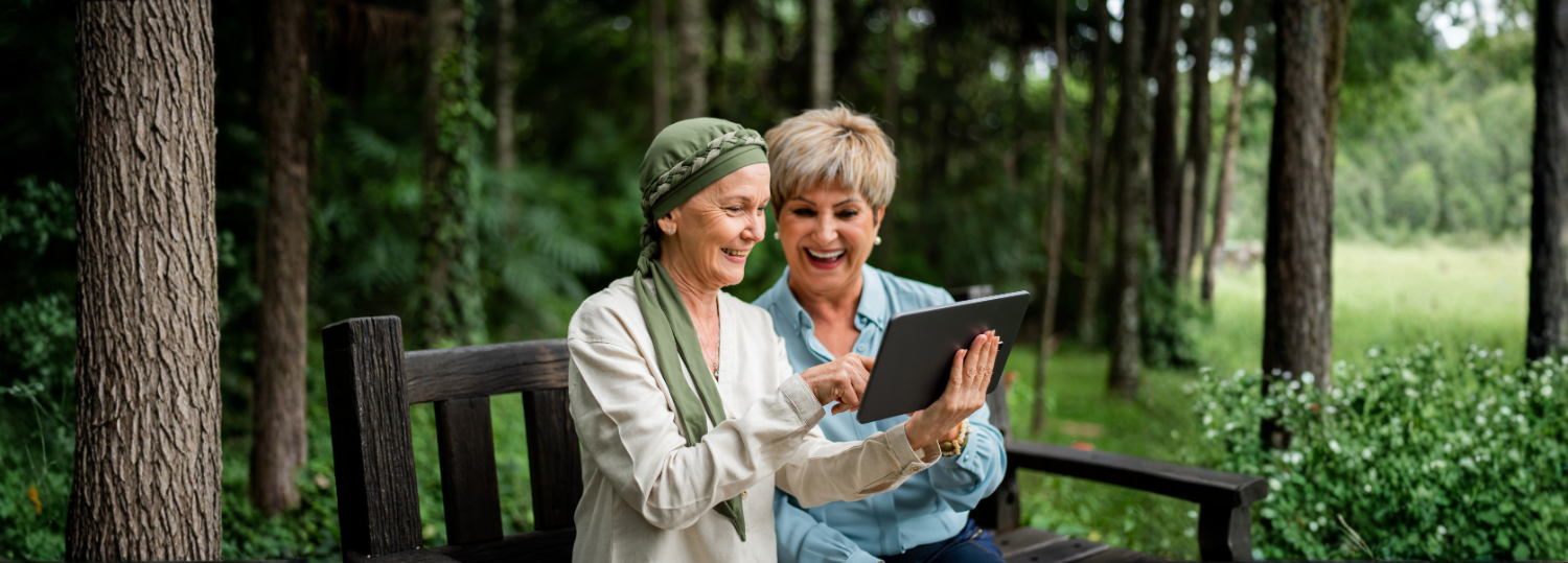 Two women on a bench, looking at a tablet. They are smiling, with a forest background.