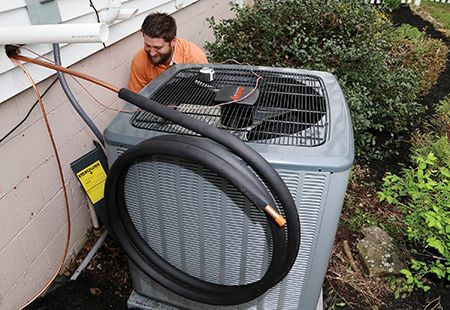 Man working on an air conditioning unit, brown tubing, near house, black insulation, exterior setting.