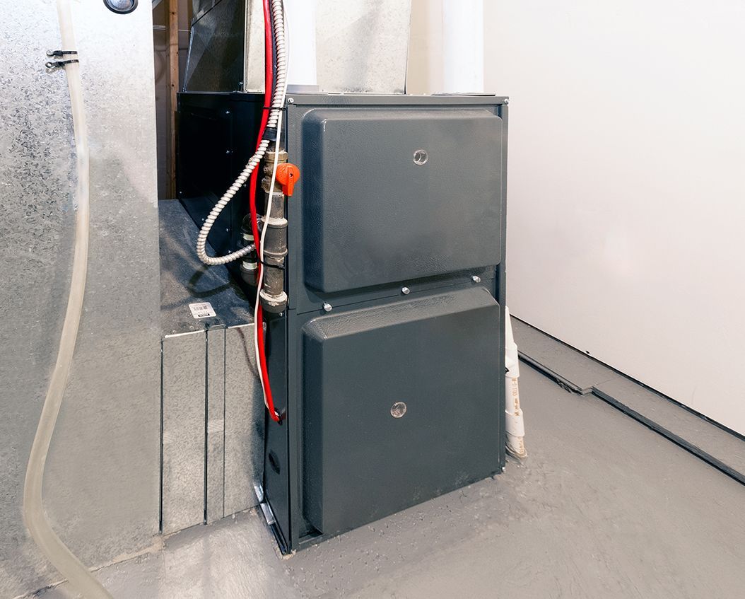 A grey industrial furnace cabinet stands on a floor next to a metallic duct, with red wiring visible on its side.