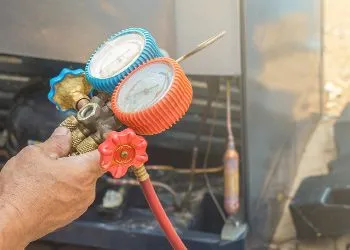 Hand holding a brass hose manifold with red and blue pressure gauges attached to a red hose outdoors