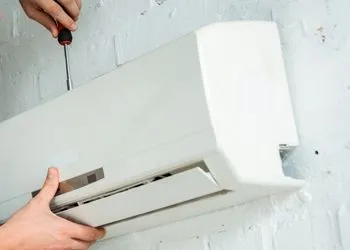 Person cleaning a wall-mounted air conditioner with a screwdriver and cloth, white indoor setting