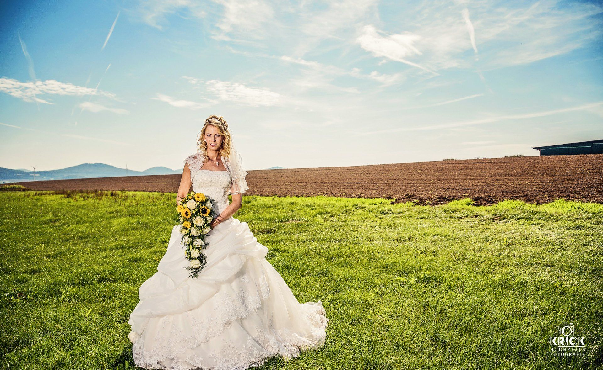 Eine Braut im Brautkleid steht auf einem Feld und hält einen Blumenstrauß in der Hand.