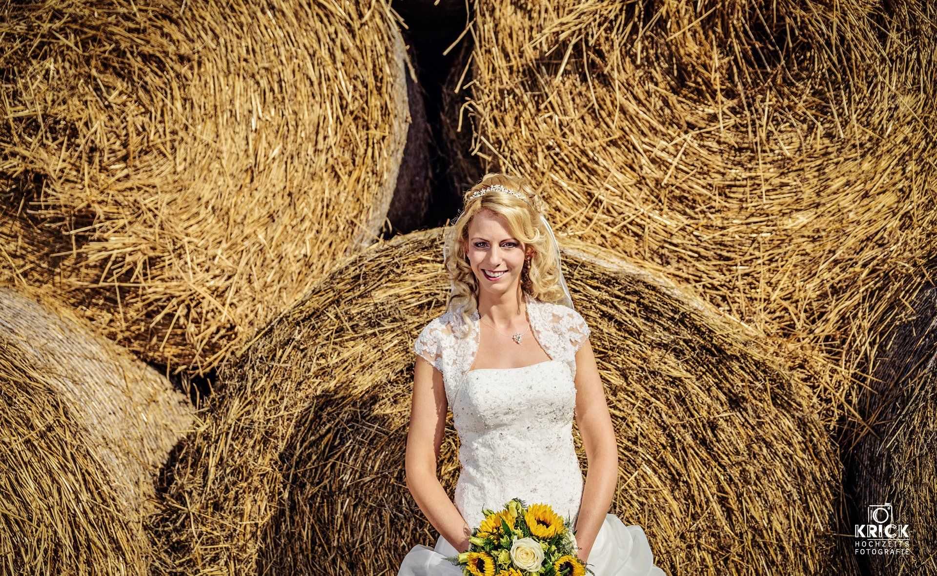 Eine Braut im Brautkleid hält einen Strauß Sonnenblumen vor Heuballen.