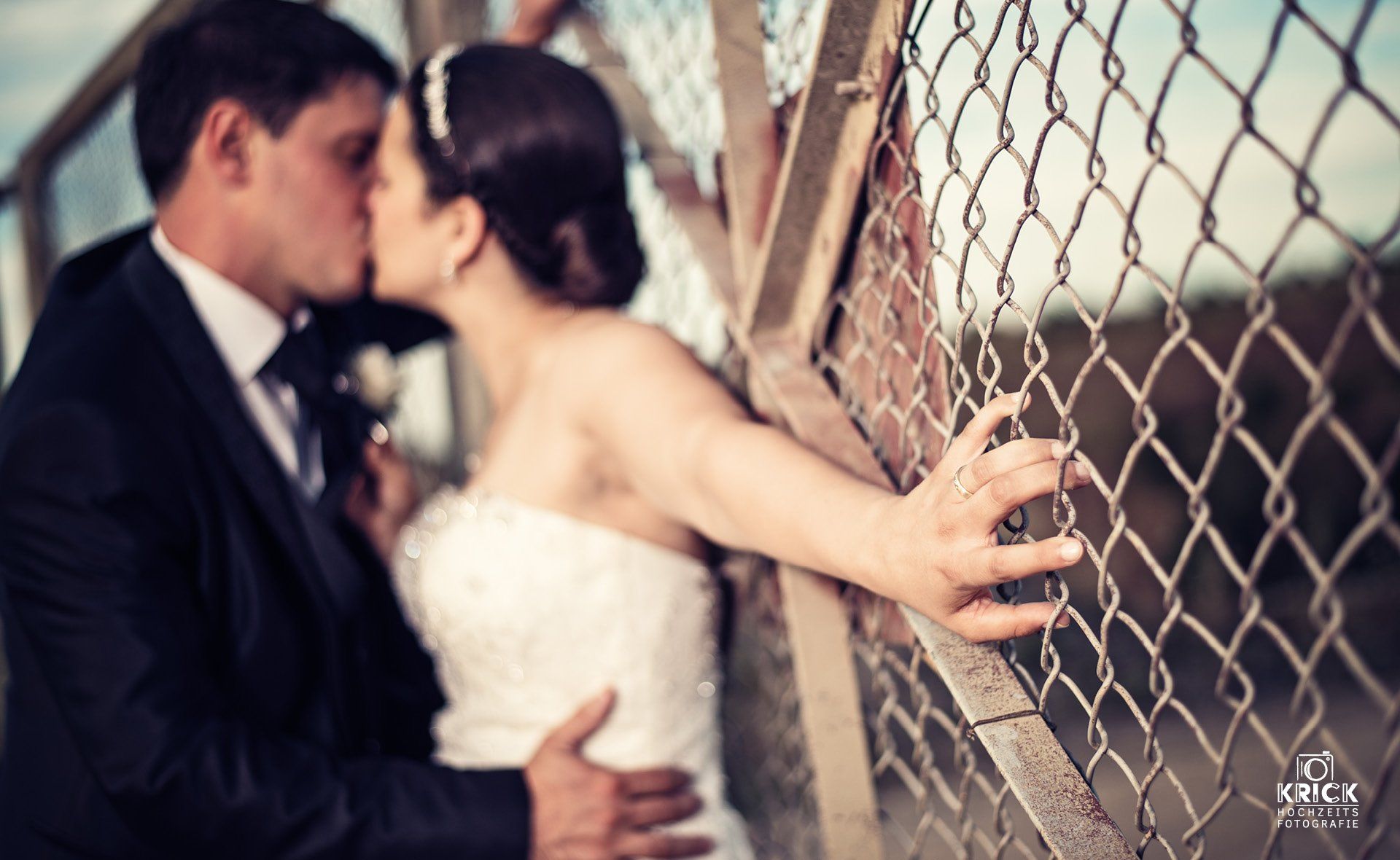 Eine Braut und ein Bräutigam küssen sich vor einem Maschendrahtzaun.