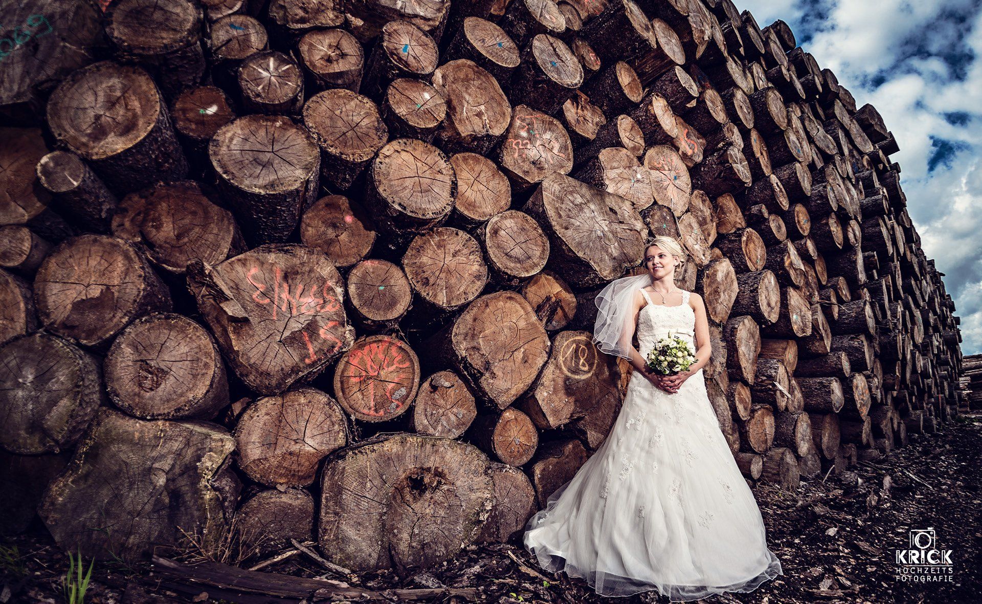 Eine Braut im Brautkleid steht vor einem Stapel Holzscheite.