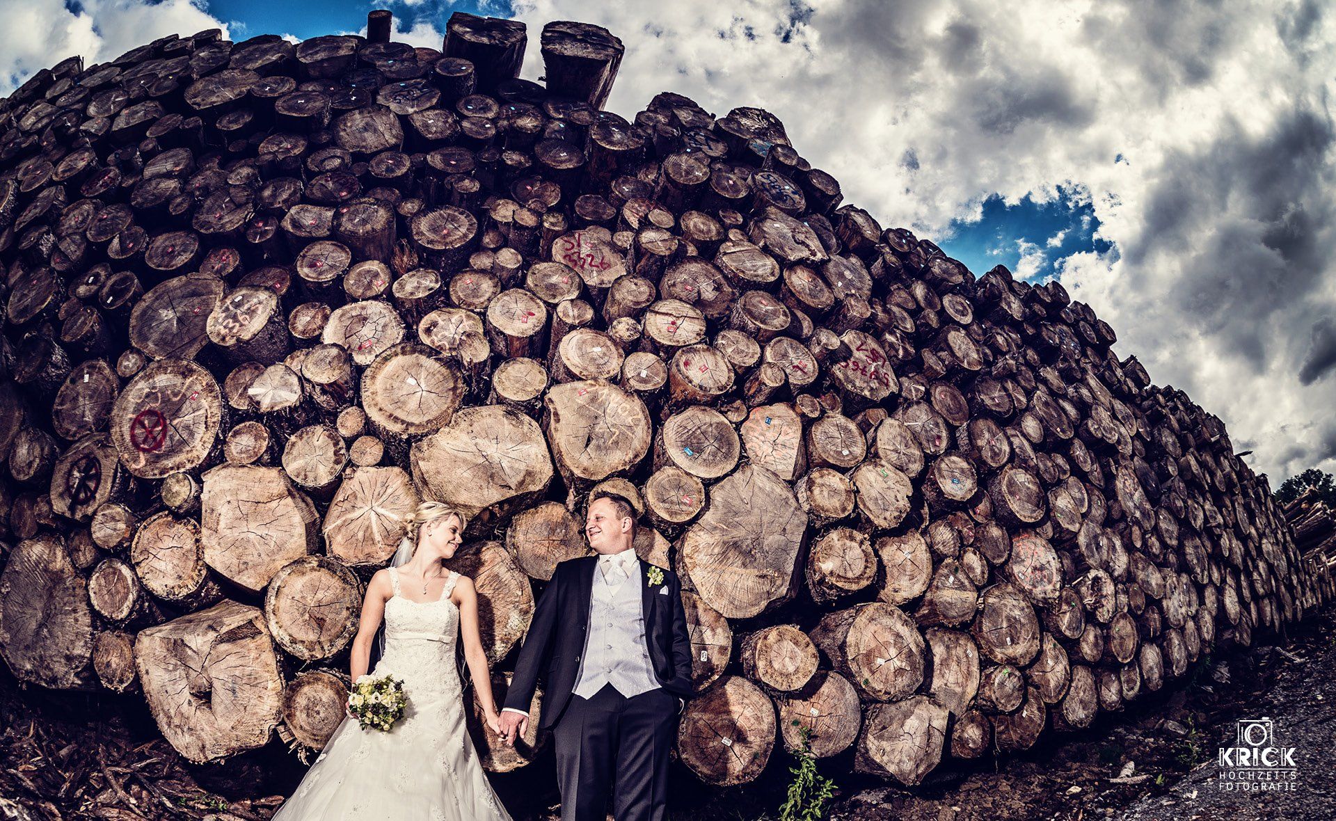 Eine Braut und ein Bräutigam halten vor einem Stapel Holzscheite Händchen.