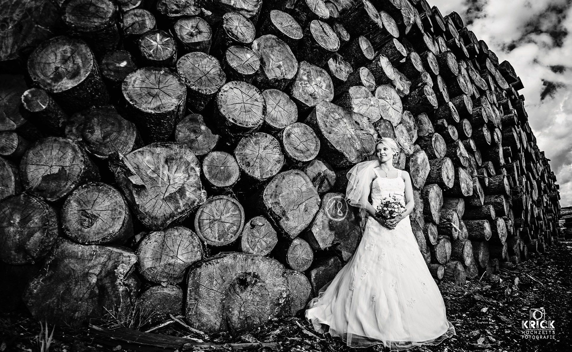 Eine Braut im Brautkleid steht vor einem Stapel Holzscheite.