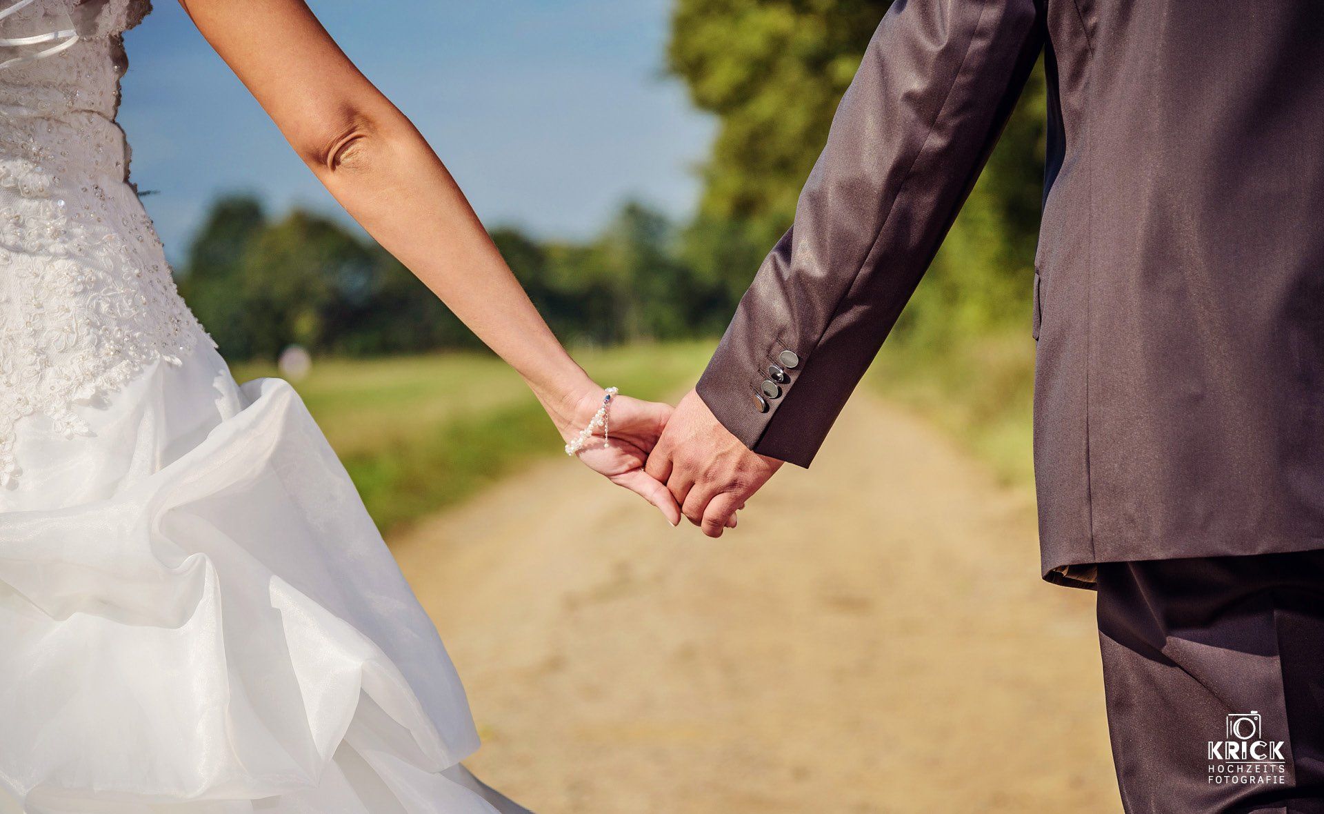 Eine Braut und ein Bräutigam halten auf einem Feldweg Händchen.