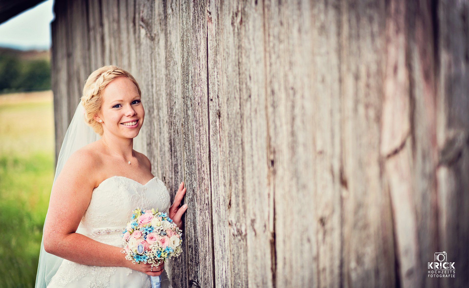 Eine Braut lehnt mit einem Blumenstrauß in der Hand an einer Holzwand.