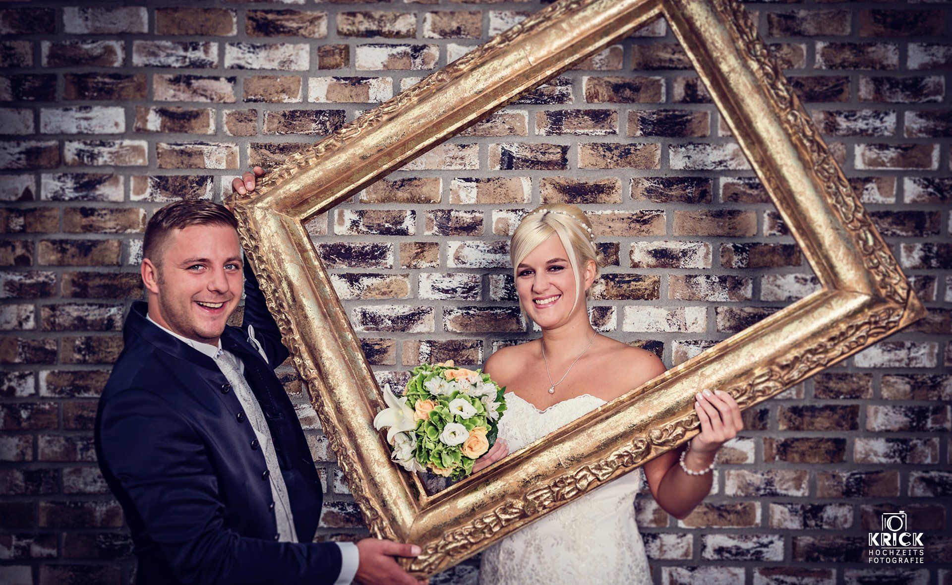 Eine Braut und ein Bräutigam halten einen Bilderrahmen vor einer Backsteinmauer.