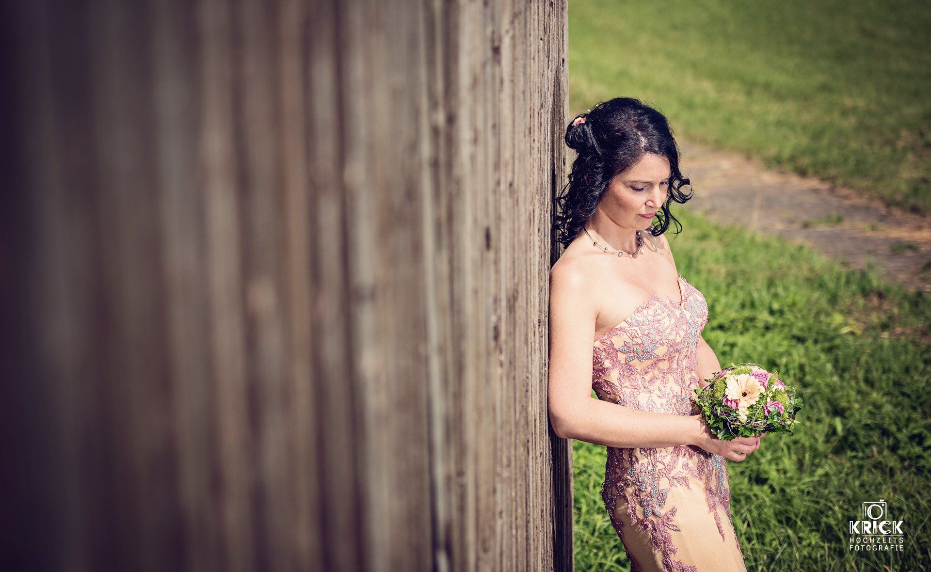 Eine Frau in einem rosa Kleid lehnt an einem Holzzaun und hält einen Blumenstrauß in der Hand.