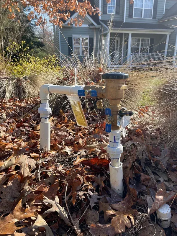 Irrigation system with pipes and valves in front yard, covered in fallen leaves, house in background.