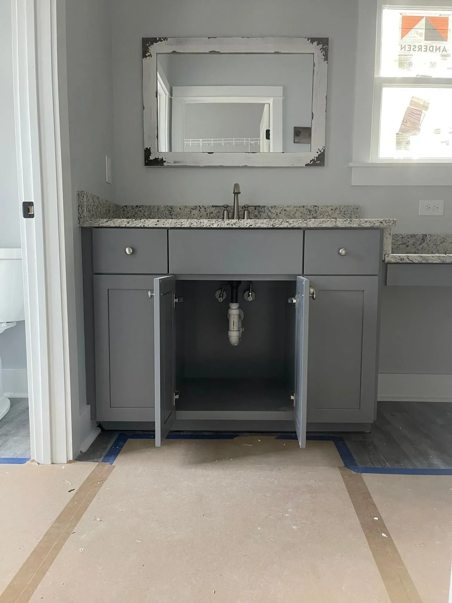 Gray bathroom vanity with open doors, mirror, and window in a new construction space.