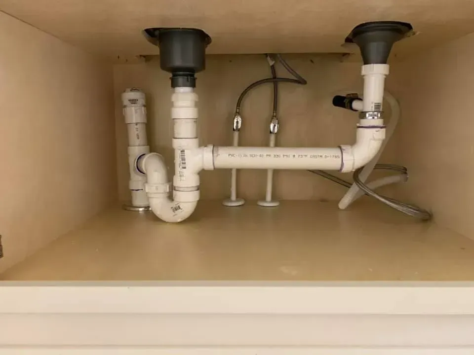 Underside of a sink cabinet with white plumbing, including pipes, drains, and a curved drain pipe.