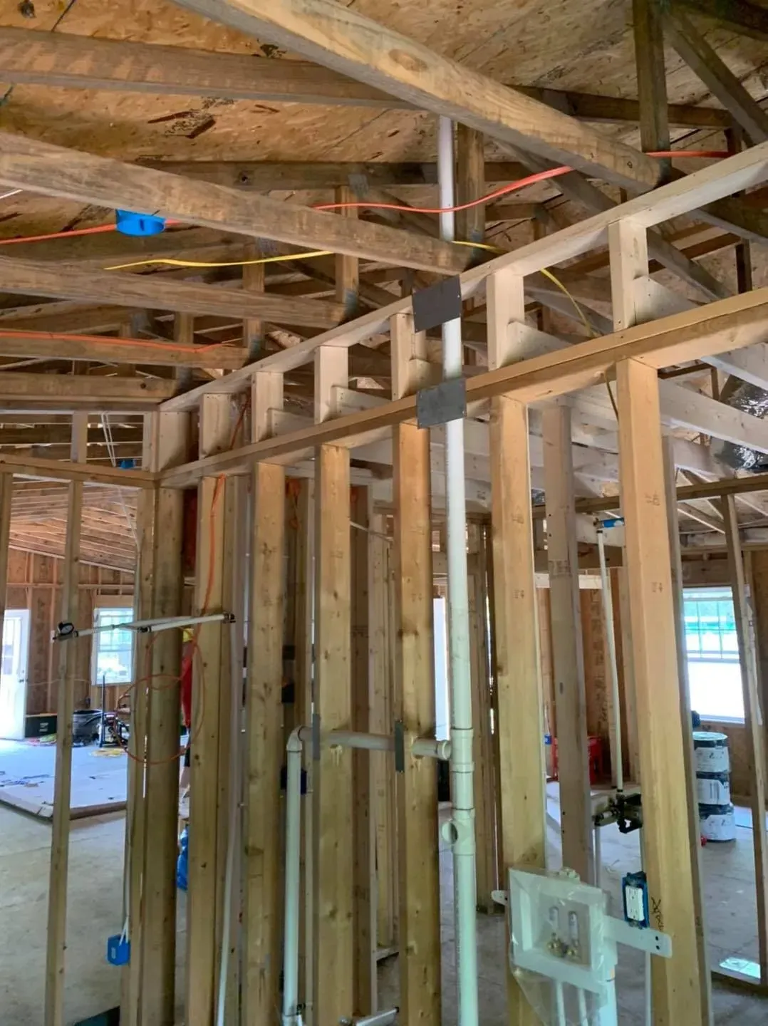 Interior of a wood-framed house under construction; plumbing and electrical wiring visible.