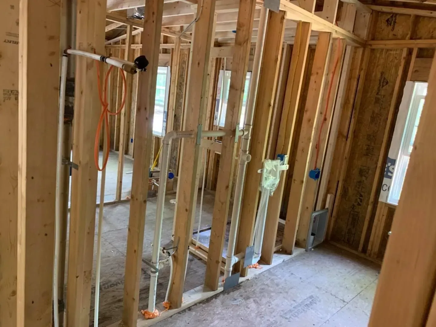 Interior view of a house under construction, showing wooden framing, electrical and plumbing, and a window.