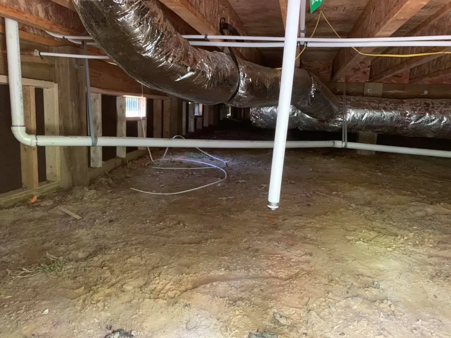 Crawlspace with exposed ductwork, pipes, and dirt floor under a house.