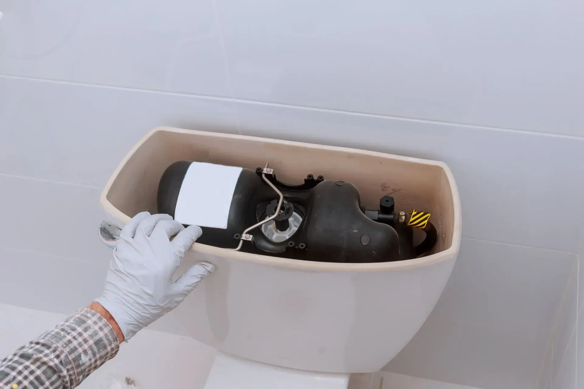 Person wearing gloves removes a black object from a toilet tank, against a white tile background.