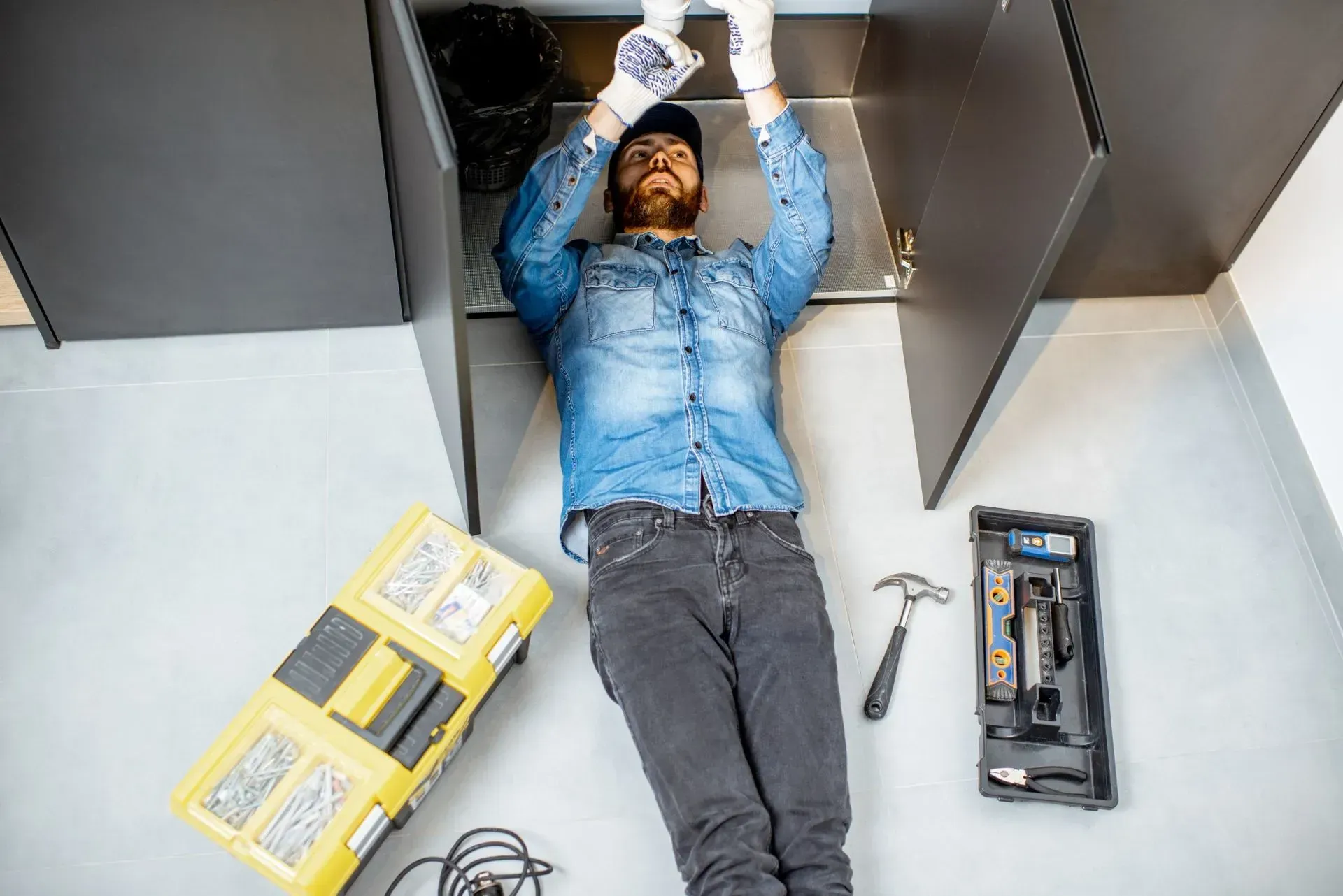 Man in jeans and denim shirt, working under black cabinets. Tools and toolbox on the floor.