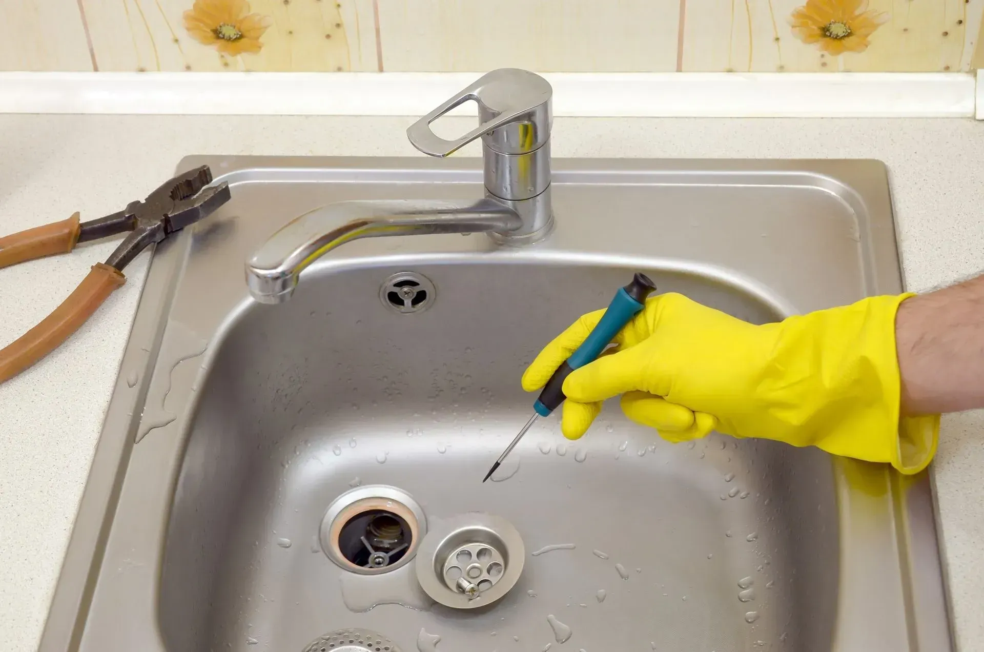 Person with yellow gloves repairs kitchen sink with screwdriver and pliers.