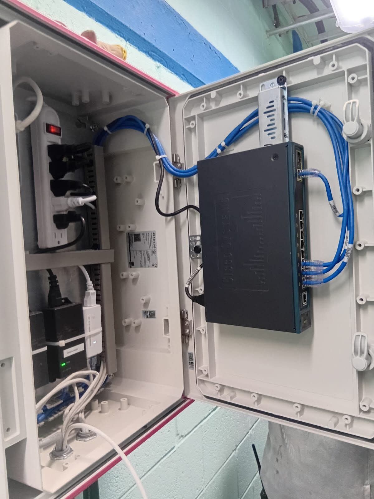 Equipo de red dentro de una caja exterior blanca y abierta. Se ven cables azules, una regleta y un router negro.
