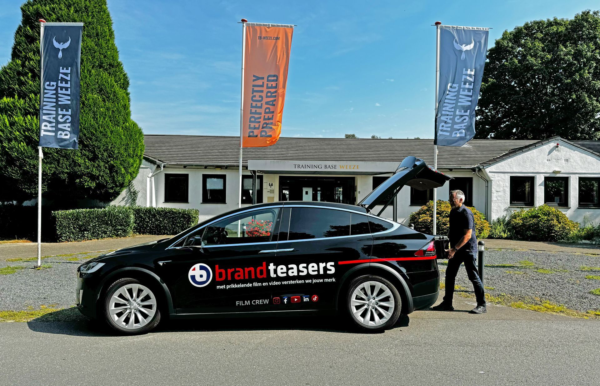 Zwarte Tesla met Brand Teasers-logo, man bij open kofferbak, gebouw met vlaggen op de achtergrond.