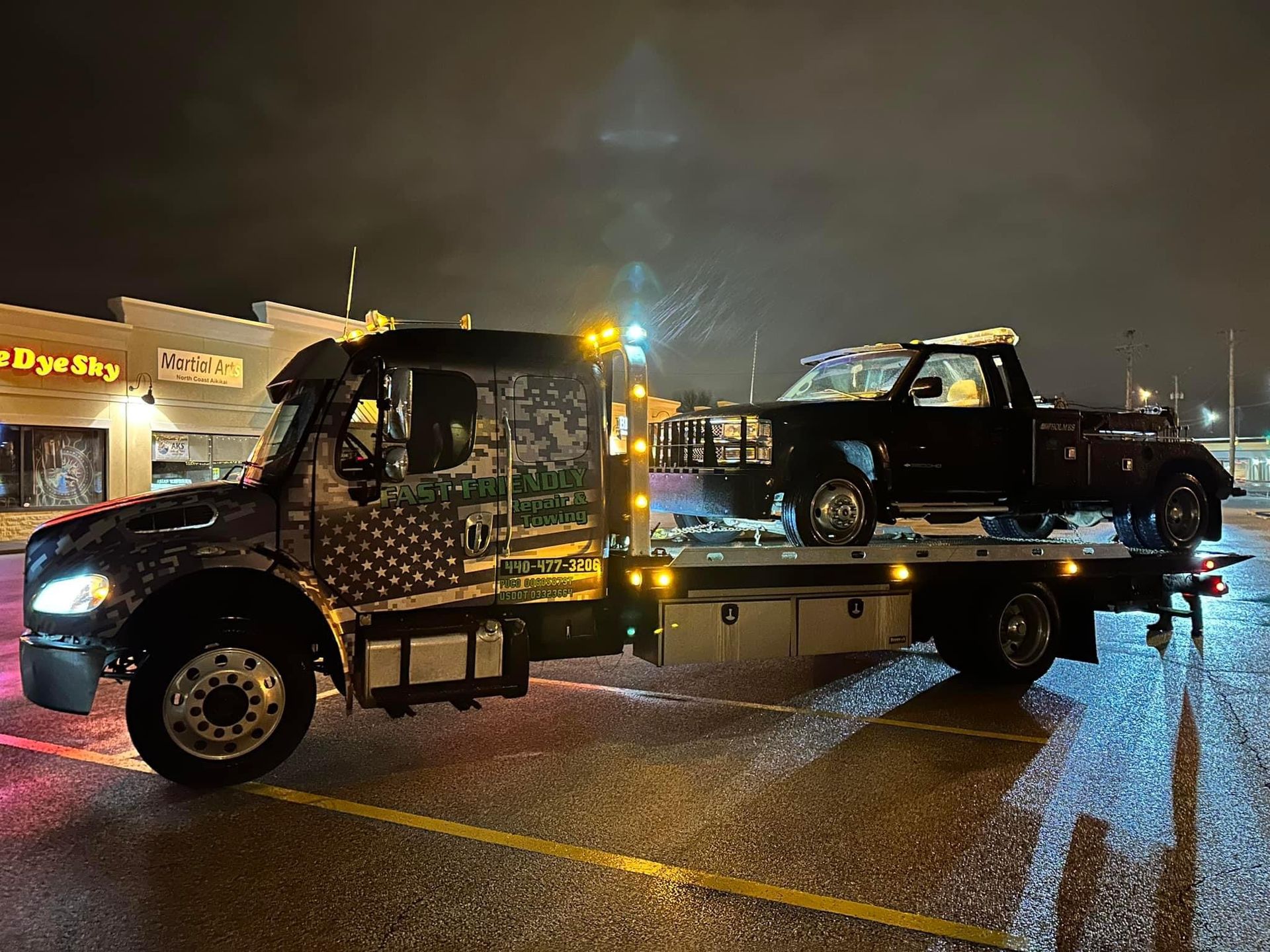 A tow truck is towing a car in a parking lot at night.