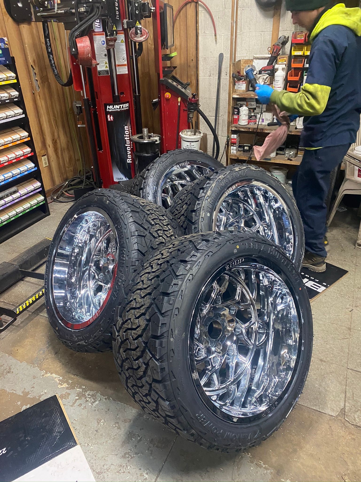 Four chrome wheels with tires in a garage; a person stands nearby.
