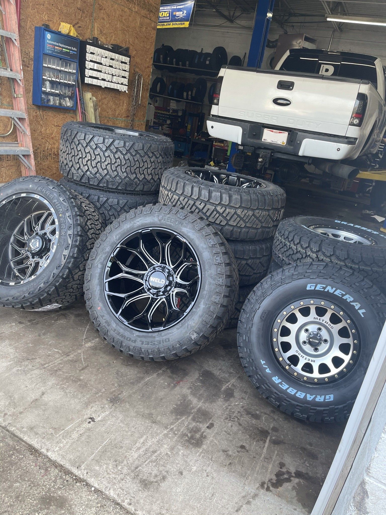 Tires and rims stacked in a garage; a white truck is in the background.