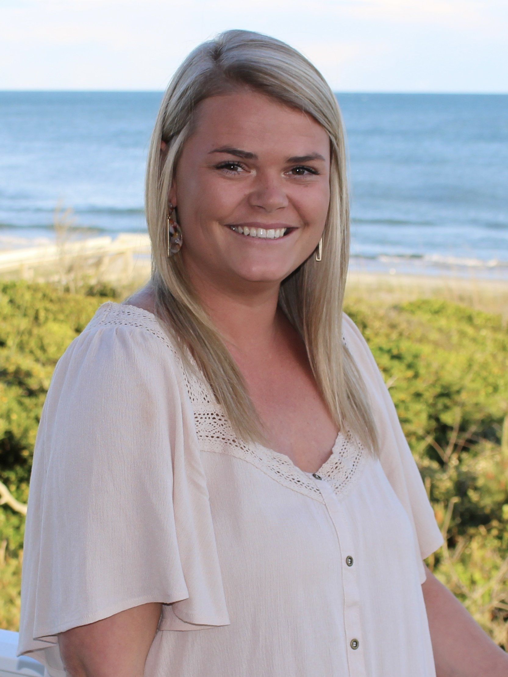 Woman with blonde hair smiling, button-down shirt, beach background.