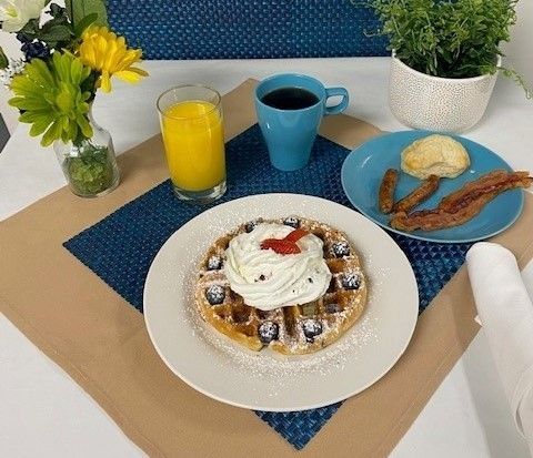 A waffle with whipped cream and strawberries is on a plate next to a cup of coffee and a glass of orange juice.