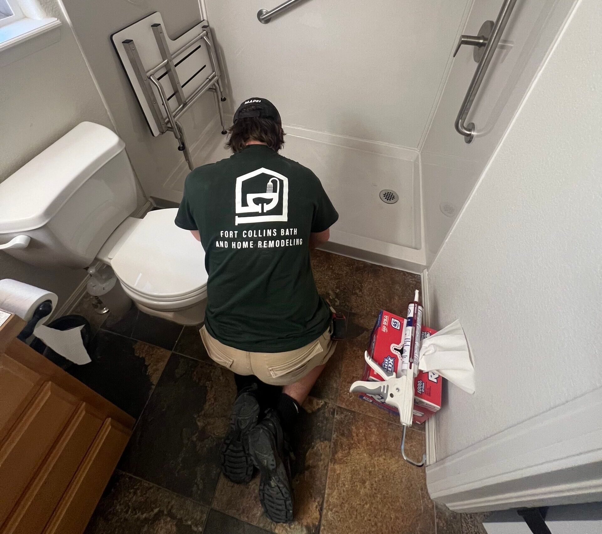 Person kneeling in a bathroom, working near a toilet and shower, with cleaning supplies on the floor