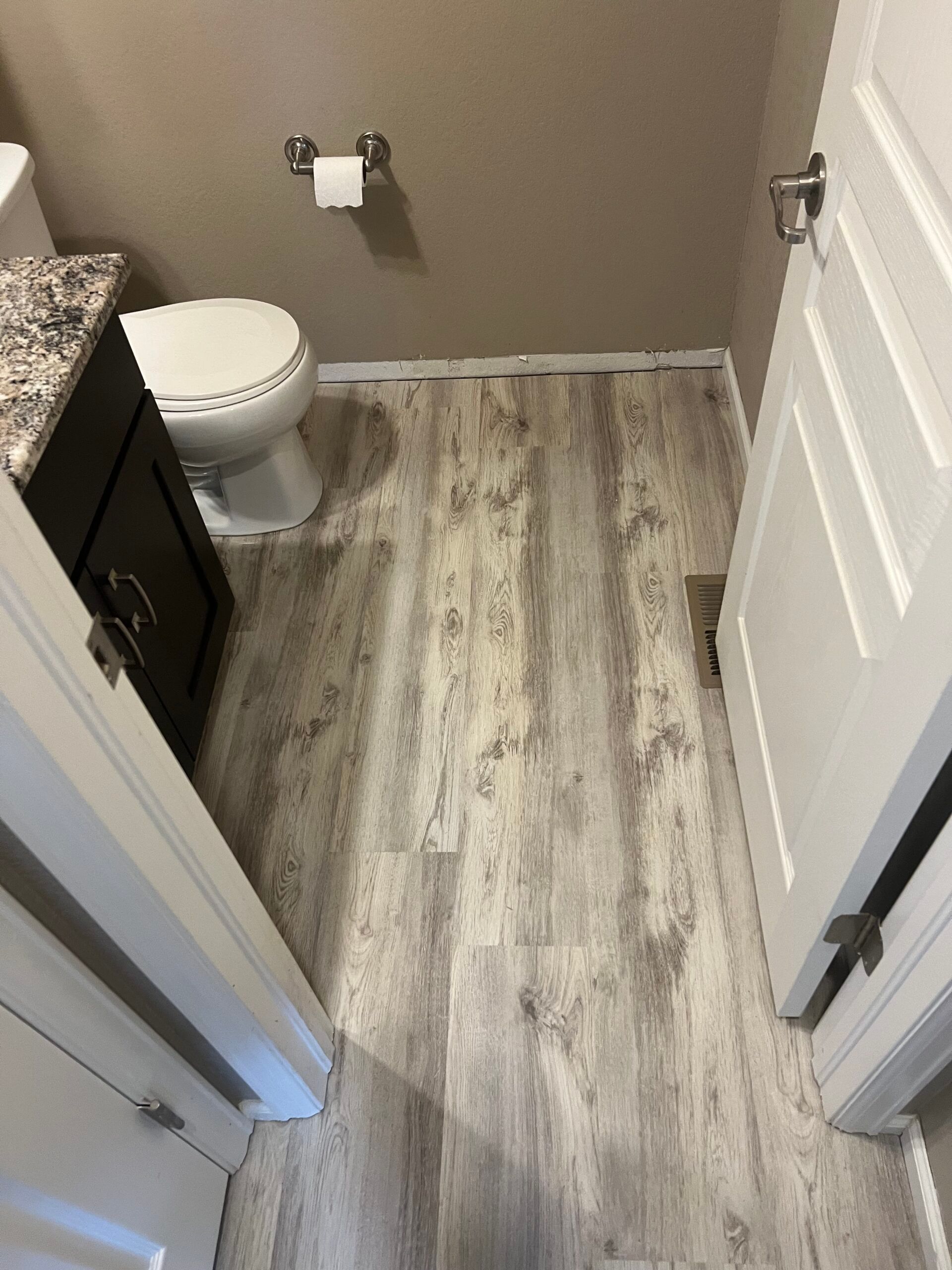 Small bathroom with gray wood-look flooring, white toilet, dark vanity, and open door.