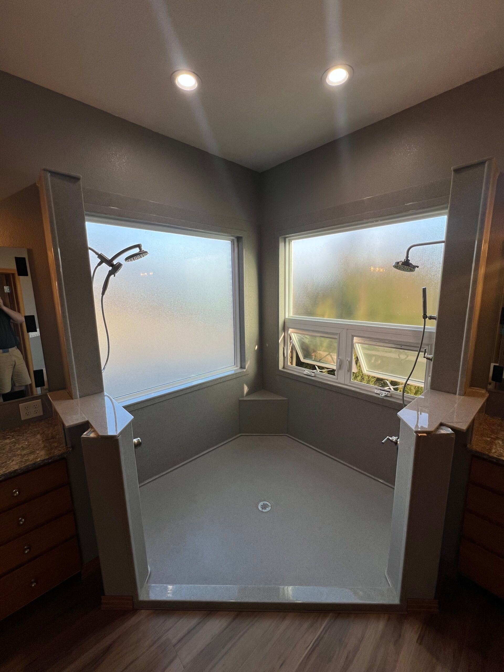 Empty bathroom with freestanding tub, gray walls, frosted window, and two corner showers under recessed lights