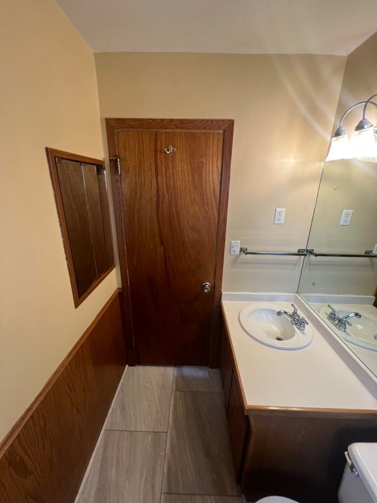Small bathroom with wood door, white sink vanity, mirror, and beige walls.