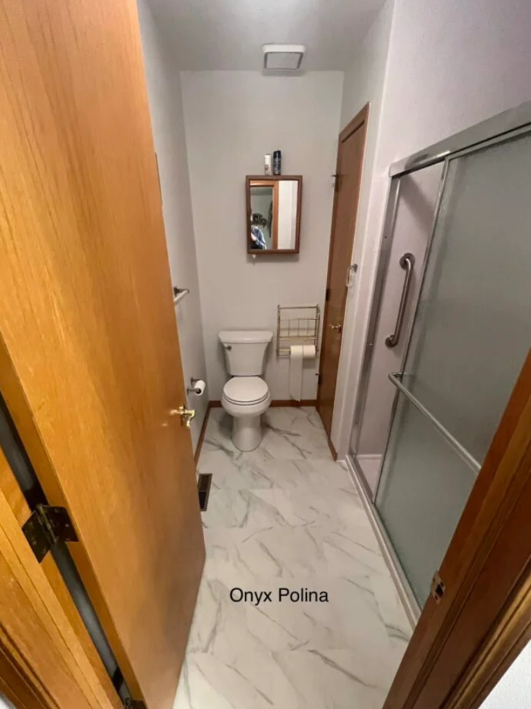 Narrow bathroom with white marble floor, toilet, sink, mirror, and frosted shower door.