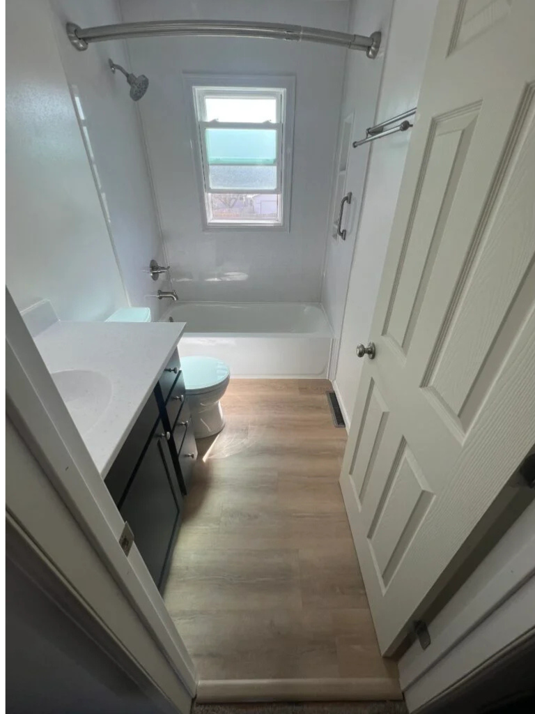 Small bathroom with white tub, window, toilet, and wood-look floor seen from the doorway