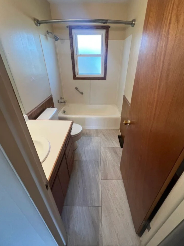 Narrow bathroom with bathtub, toilet, sink vanity, wood door, and window with curtain rod.