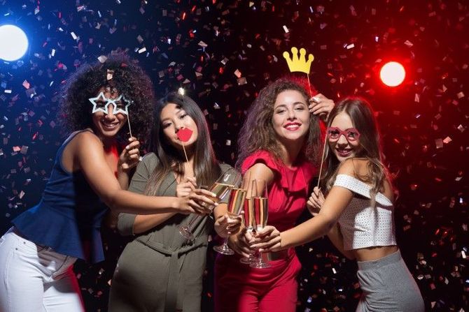 Four friends in party hats pose with confetti and colorful lights, smiling and holding drinks.