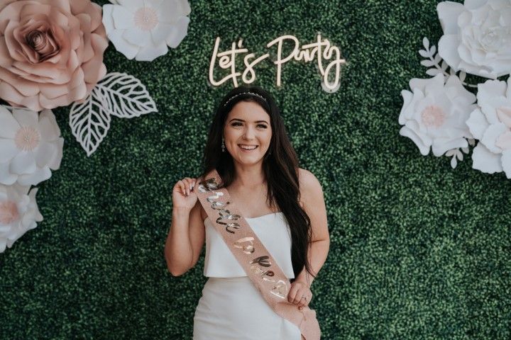 Woman in white dress and sash posing before a greenery wall with large flowers and a “Let’s Party” sign